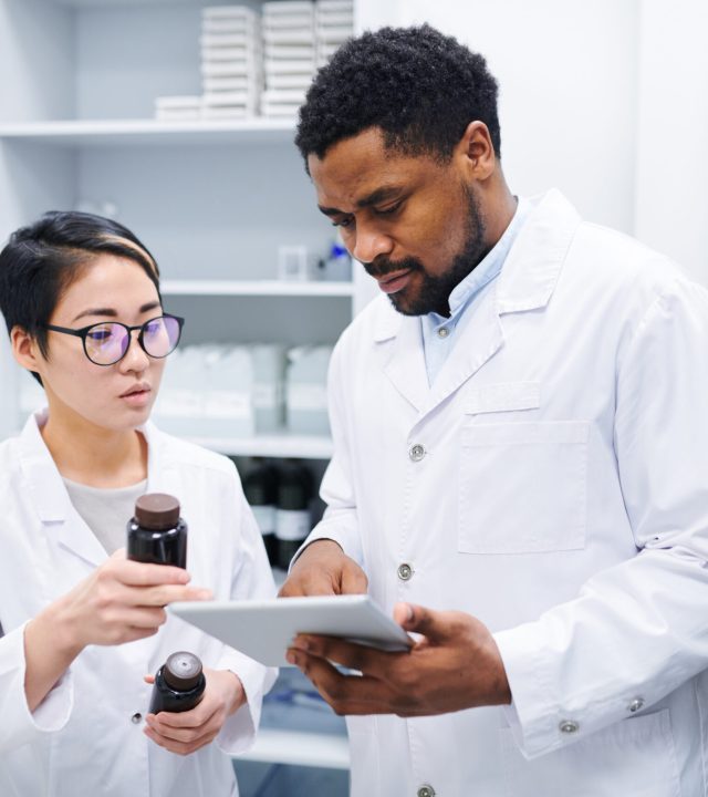Serious thoughtful multi-ethnic pharmacists in lab coats standing in drugstore and discussing new medications while using tablet to search for information in database